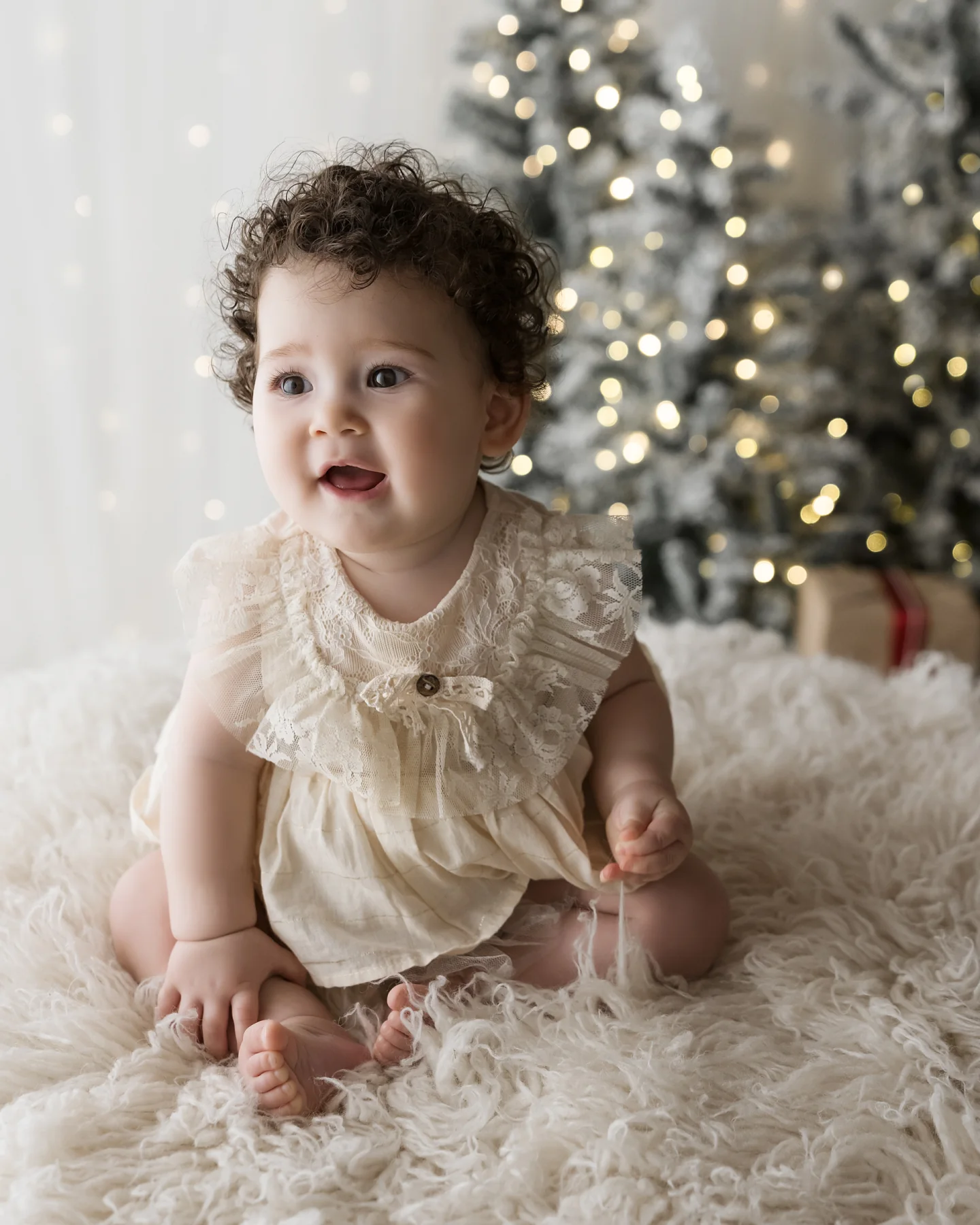 Bambina riccia con il vestito color panna seduta sulla pelliccia davanti all’albero